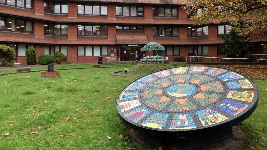 In the foreground is a circular multi coloured mosaic on a green space. In the background there is the Surrey Heath Borough Council offices in Camberley. 