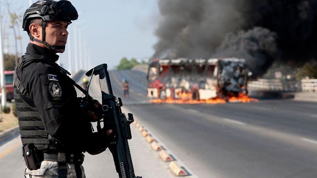 An armed Mexican police officer looks on while a bus burns on a highway in the background