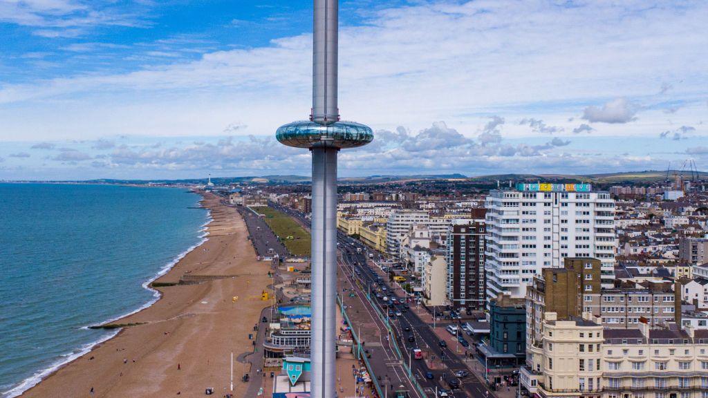 The i360 tower up in the air with Brighton's beach and seafront in the background on a bright day.