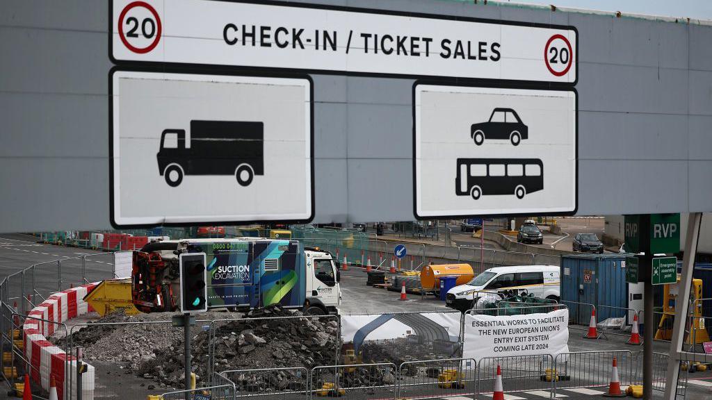 A sign which reads check-in/ticket sales and has pictures of cars and lorries to show which lanes to use. Underneath and in the background you can see construction work going on with a pile of rubble, skips and machinery around