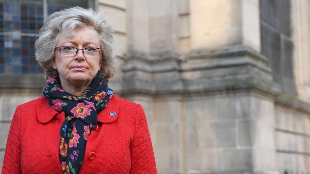 A woman in a red coat and floral scarf standing in front of a Birmingham's sandstone cathedral building. A stained glass window can be seen in the background. The woman has greying blonde hair and is wearing dark framed glasses. She has a serious expression.