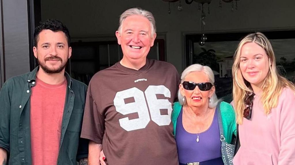 Four people are stood next to each other. From left to right - a man with beard and moustache and dark hair is wearing a red t-shirt and green shirt. Next to him is an older man with grey hair, wearing a brown t-shirt with the number 96 on it in white. Next to him is an older woman with long white hair, wearing sunglasses. She is also wearing a purple top with belt and green cardigan. At the end is a young woman with long blonde hair and pink top with sunglasses hanging from the collar.