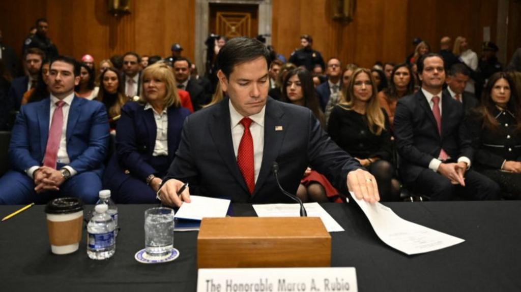 Marco Rubio is surrounded by people as he adjusts some papers at a desk