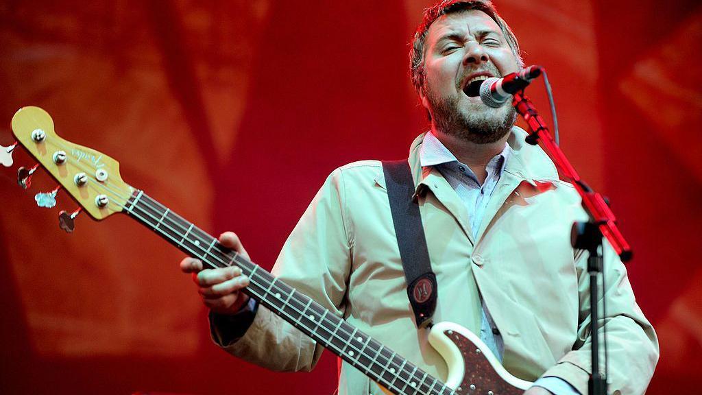 Jimi Goodwin, with short hair and a beard, wears a pale cream jacket and white shirt as he plays a guitar and sings into a microphone in front of a red background