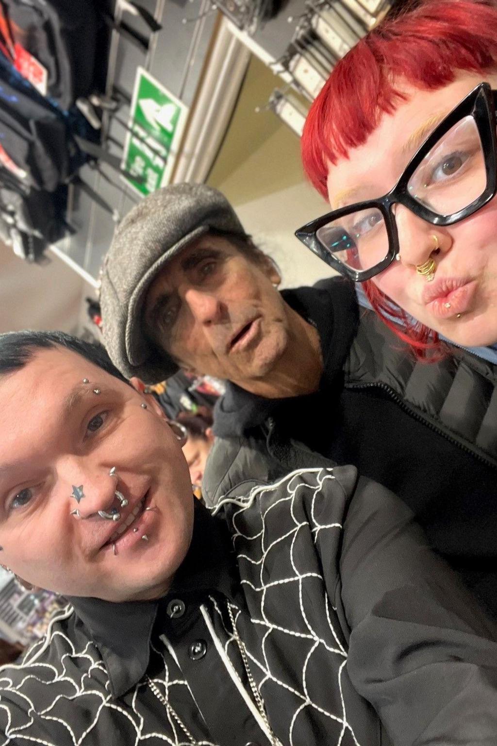 Adhum Price, a man with bright green and black hair and numerous facial piercings, is posing for a selfie alongside Alice Cooper who is wearing a tweed flat cap, and stood in between Mr Price and a pouting store colleague who has bright red hair and is wearing black, thick-rimmed glasses.