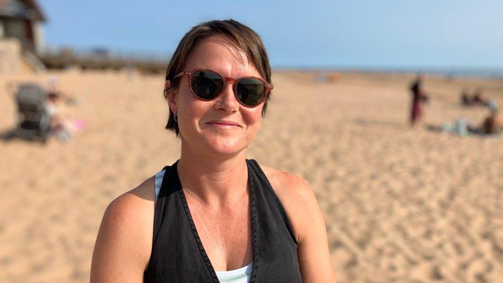 A woman sitting on the beach and smiling at the camera. She is wearing a black halter neck top and sunglasses. A sandy beach is in the background.