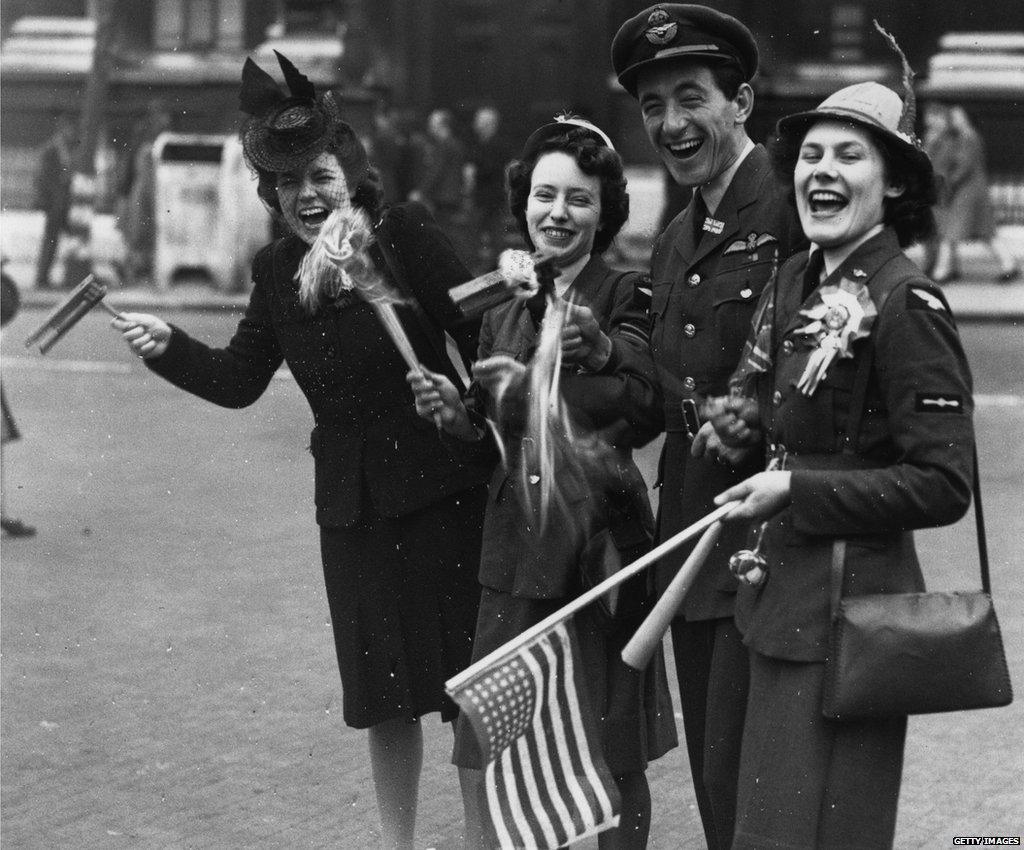 A RAF officer, two members of the Women's Royal Airforce and a civilian celebrate in London's Whitehall. One person holds and American flag.