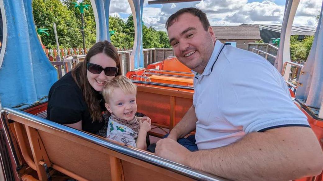 A smiling young family sitting together on a roller coaster rider. The man in a white polo shirt is on the right, a woman with long dark hair and shades sits on the right and a smiley blonde boy sits in the middle.
