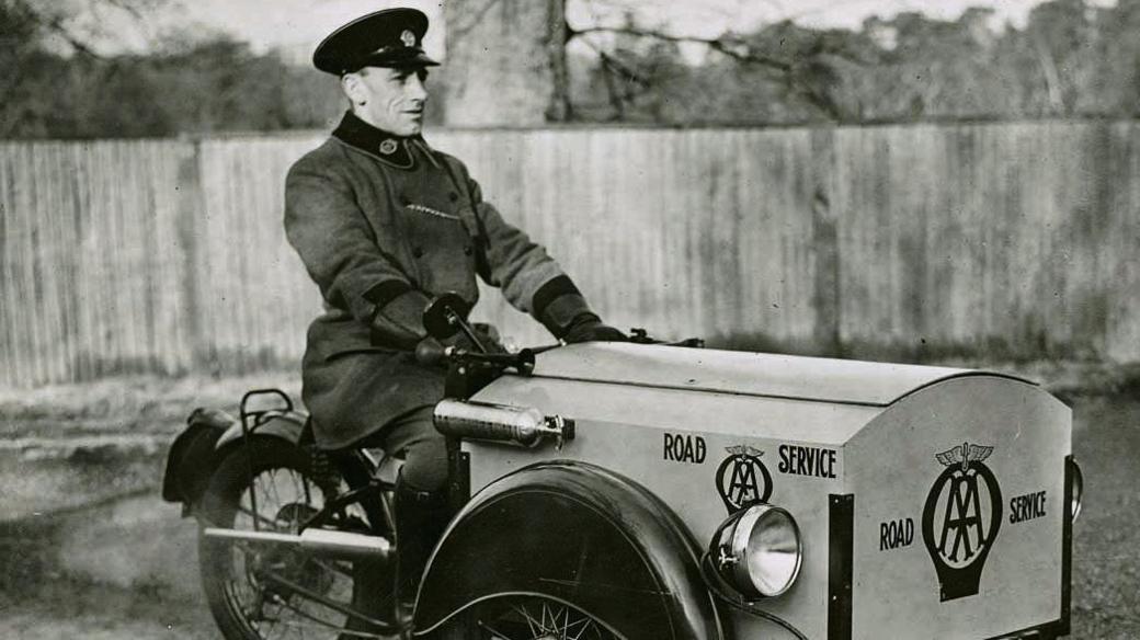 A black-and-white photo of a three-wheeled motorcycle bear the AA logo and being driven by a man in uniform.