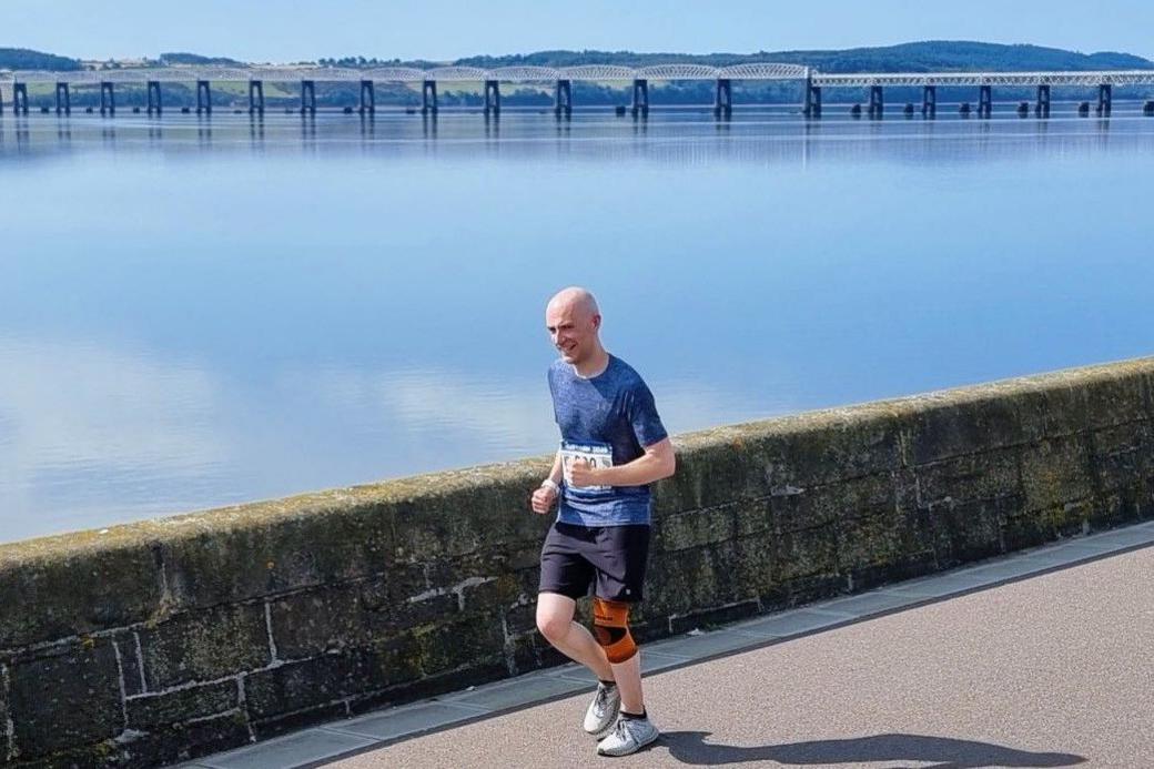 Thomas Mills runs along the still River Tay wearing a blue top, black shorts, white trainers and an orange knee brace. He smiles at the camera.