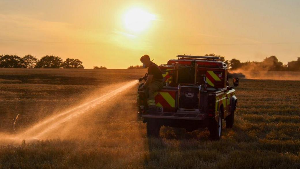 Firefighter with water spraying from a hose sat on a 4/x fire appliance in a field which is smoking. The sun is setting.