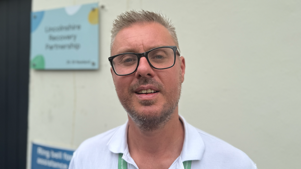 The photo shows a man looking at the camera. He's got short, spiky blonde hair. He's wearing black frame glasses, and a white top with a colour. He's standing in front of a white building.