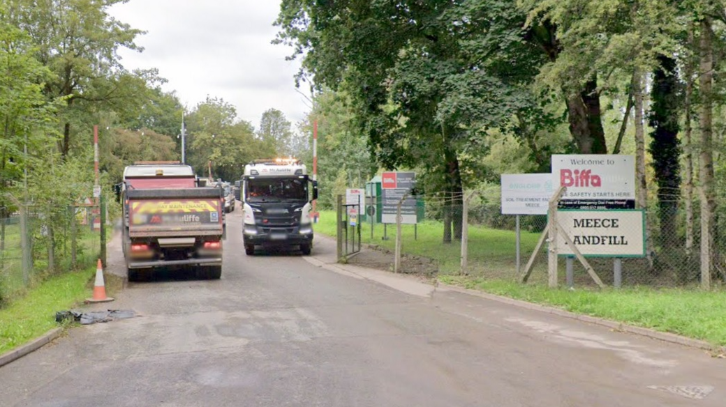 A road leads into a gated-off area in amongst trees. Two lorries are driving on the road. A nearby sign reads "Meece Landfill", while another reads, "Welcome to Biffa".