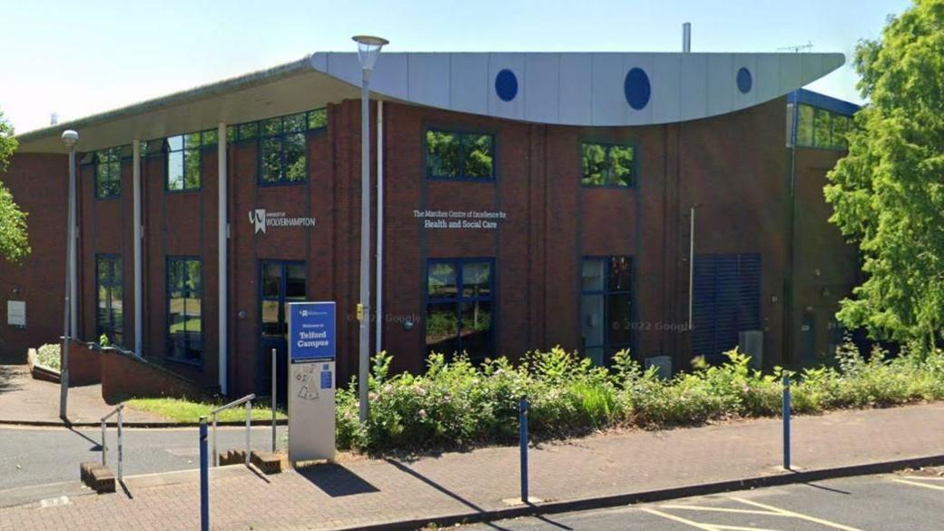 The exterior of a building at the University of Wolverhampton's Telford campus. The picture is taken from the other side of a road near the building, showing the red-brick building with University of Wolverhampton signage, and trees and bushes around the building.