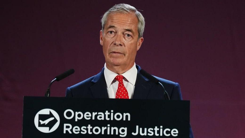 Nigel Farage, who has short grey hair parted to the side speaks at a podium with the words Operation Restoring Justice on it. He is wearing a dark suit, white shirt and red spotted tie.