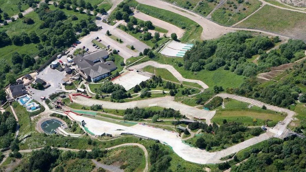 A birds eye view looking down on a dry ski slope snaking through a grass area with trees and buildings