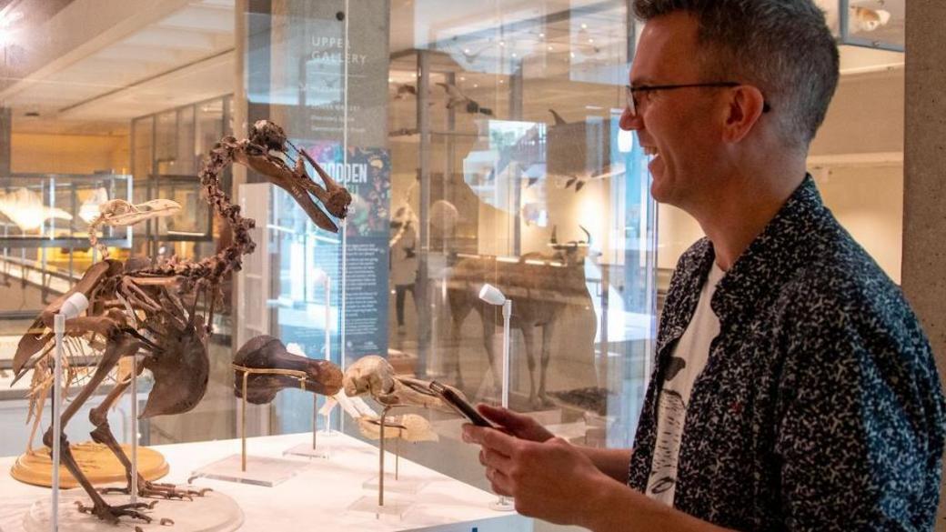 A skeleton of the extinct flightless bird the dodo in a clear glass case. Jack Ashby, in a blue and white shirt over a blue T-shirt, is standing side-on to the case, smiling towards the dodo and holding up a smartphone.