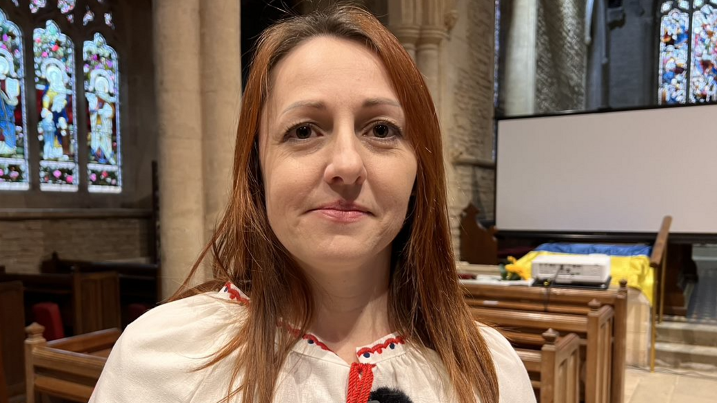 Olga Bilokin is standing in a church. She is smiling at the camera. She has brown hair dyed with a red tinge and is wearing a white top with a red pattern on the collar. Stained-glass windows and pews can be seen behind her.