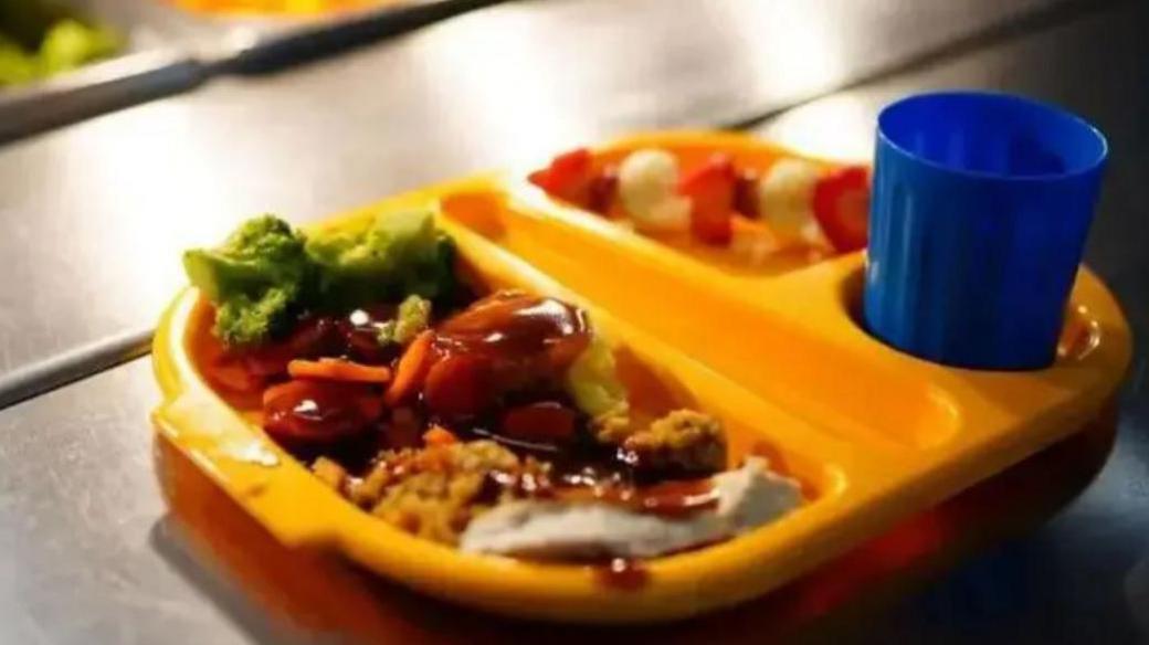 A yello compartmentalised tray containing a school meal of meat and vegetables with a blue cup