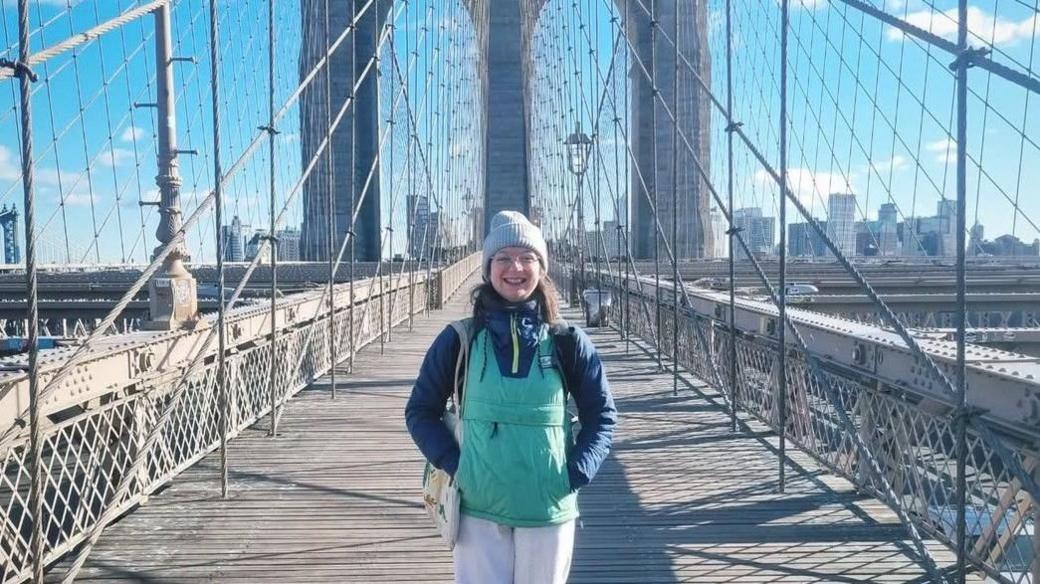 Becky in a blue woolly hat and green jacket standing on the Brooklyn Bridge in New York