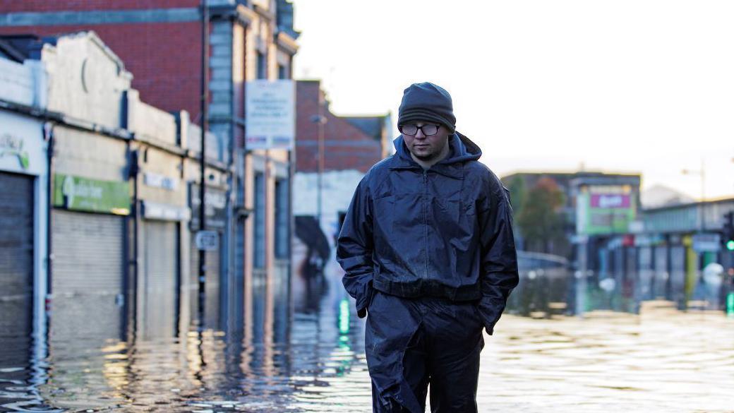 A man in waterproofs and welly boots walking through water that is up to his shins. On either side of him, shopfronts are visible, making it clear he is walking down a street