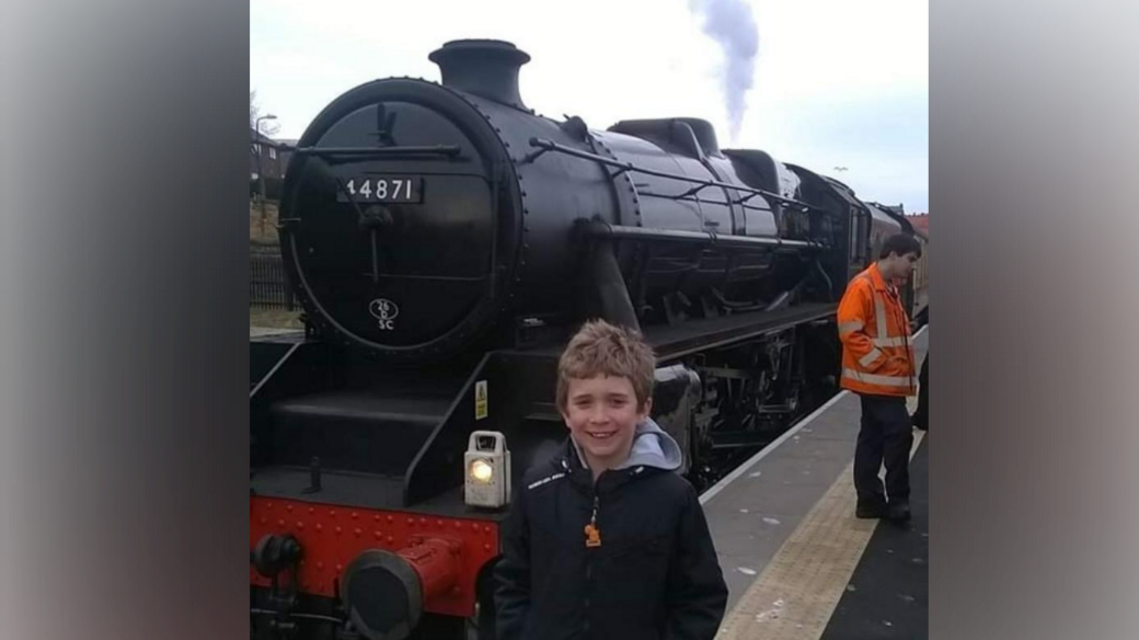Logan Smallwood pictured as a younger boy smiling as he stands in front of a black and red steam train, sitting on a platform