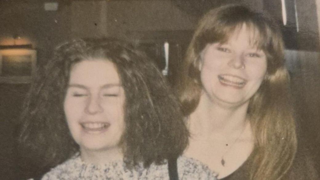 An old photo of two young women on a night out. They are grinning at the camera and standing beside each other. One has dark hair and the other has brown hair. They are standing in a darkly lit indoor venue.