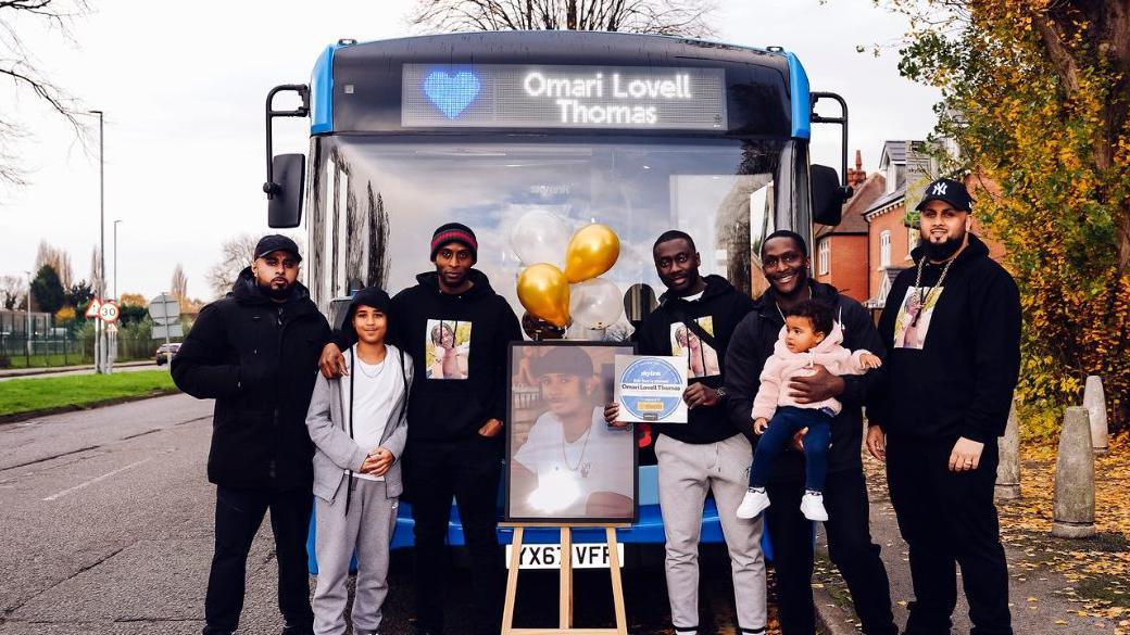 Family and friends pose with a portrait of Omari in front of the Skylink bus