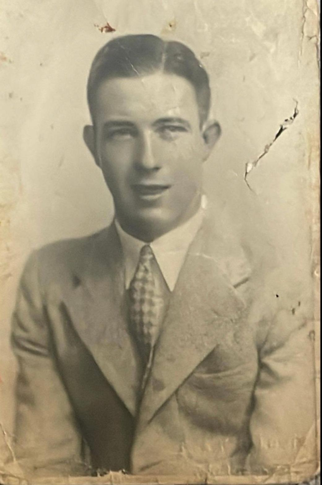 The old black and white photo shows a young man in a suit, shirt and tie looking at the camera. His dark hair is smartly slicked back. The photo is slightly torn.