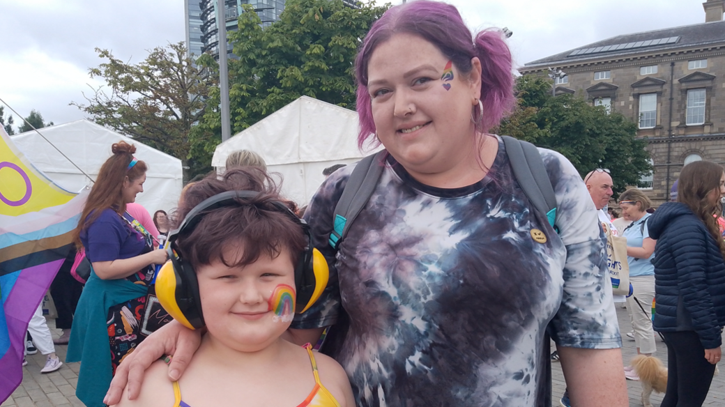 Jenna standing with her arm around Charlie's shoulder. In the background people walking about and carrying pride flags. Charlie is wearing a rainbow top, headphones, and has rainbow face-paint