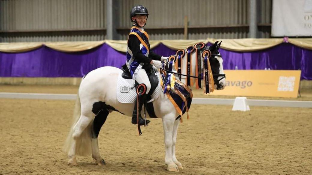 Theo wearing a black riding helmet, jacket and white jodphurs sits on George, a piebald horse, which is white with black patches. George's mane is covered in orange and purple rosettes and Theo has an orange and purple sash over his chest.