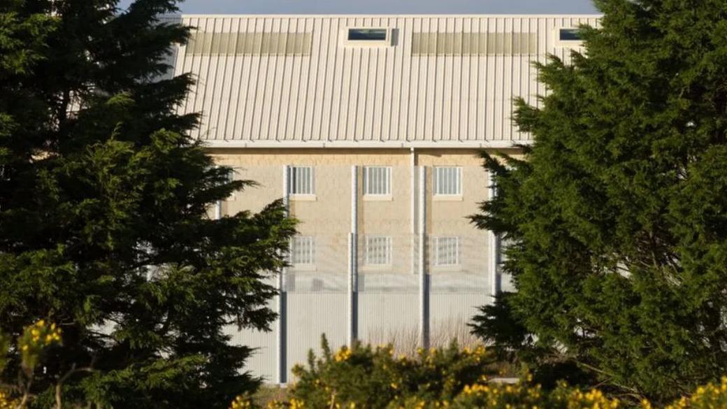 A fence and a wall of HMP La Moye is seen through some trees, with barred windows also visible