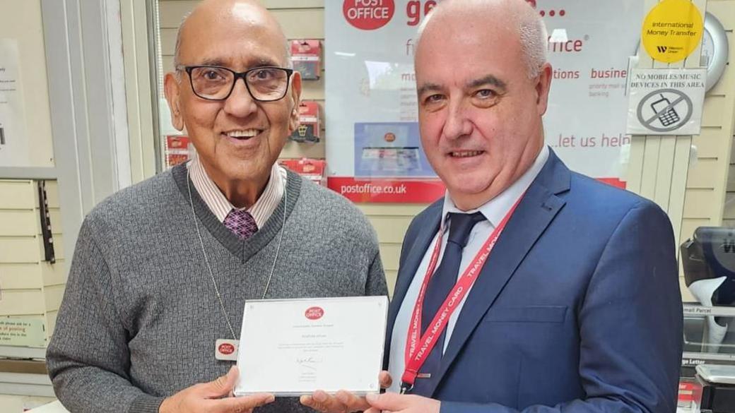 Arshad is wearing a grey jumper, shirt and tie and is holding his long service award standing next to Paul Spry who is wearing a blue suit and blue tie, in the background is the Post Office counter.