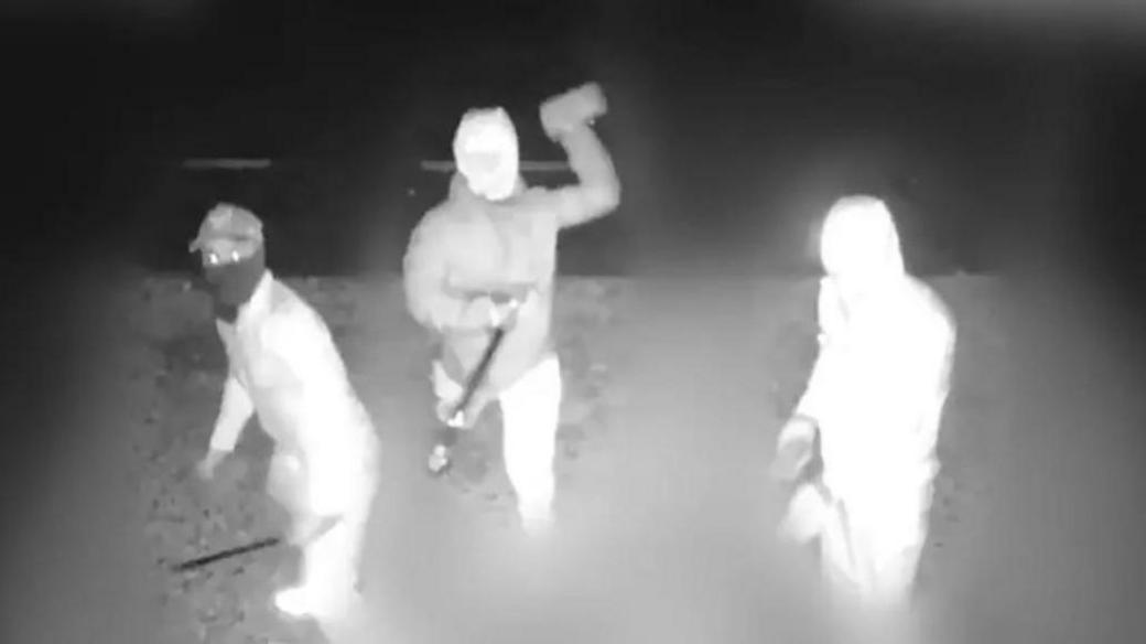 Black and white CCTV footage showing three people with face coverings, carrying machetes and throwing bricks through the windows of the property.