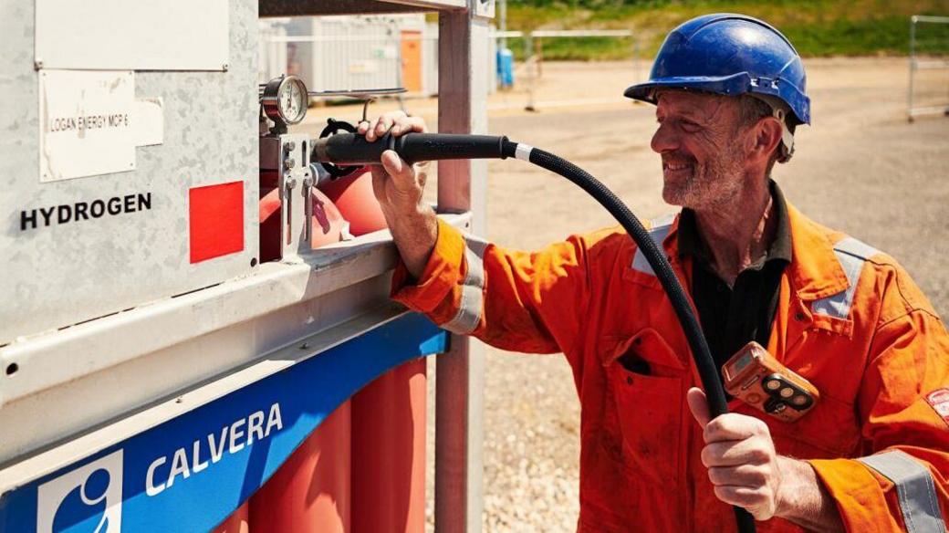 A worker at Canford Renewable Energy holds a tube connected to a hydrogen facility. He wears a blue helmet and orange overalls.