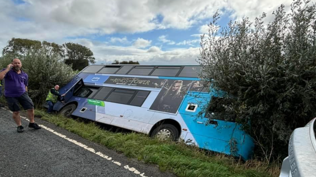 A double decker bus on its side in a ditch. The bus is blue and white. Two men are stood near it - one is by the bus door, the other is up on the road on the phone. 