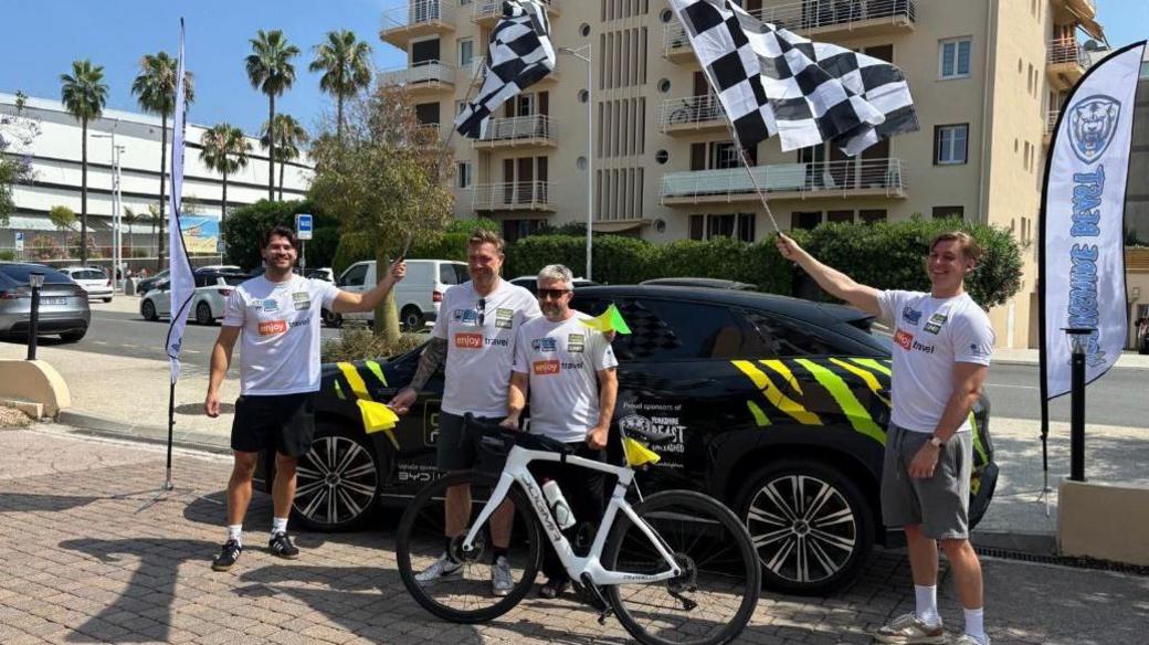 Four men stand next to a black car in Nice, some waving chequered flags and one standing next to a bike.