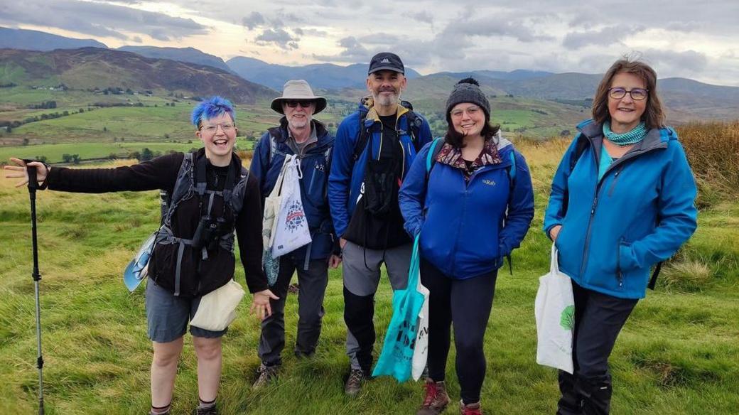 Five volunteers dressed in outdoor clothing pose for a photo near the mountain. They are all carrying bags for collecting seeds.