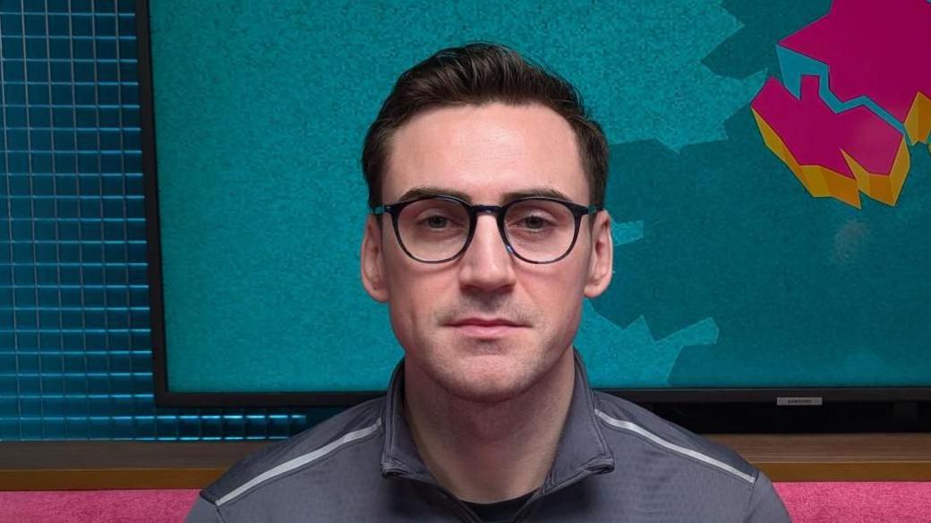 A man is looking straight at the camera. He has short jet black hair, which is slicked back. He has black-rimmed glasses. He is wearing a grey sports top with a zip. He is sitting in a podcast studio with a pink sofa, behind his head is a TV with the logo for The State of Us podcast, which is a teal background with a stylised image of Northern Ireland in pink.