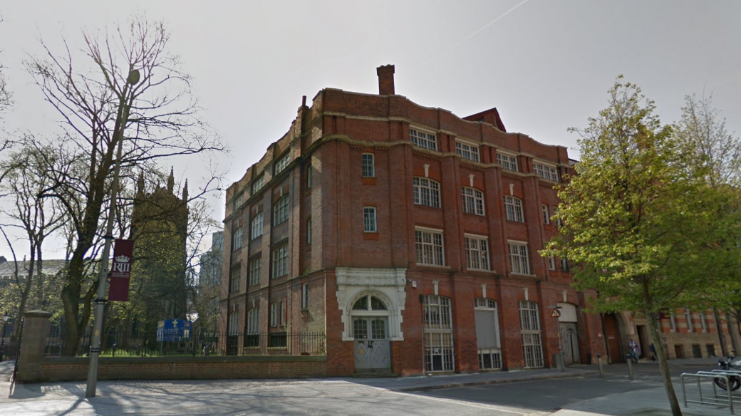 An imposing four-storey red brick building on a tree-lined street next to a church