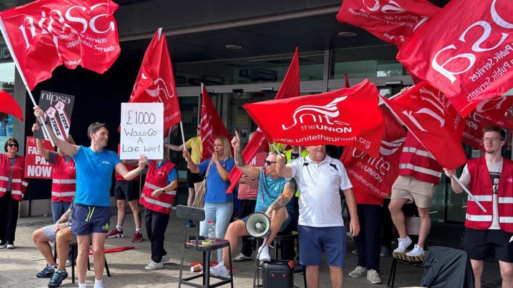A group of people from Unite the Union on strike at the door of a leisure centre. They are wearing different coloured clothing and holding large red flags and a poster with the 'unite the union' and '£1000 would go a long way' written on them. There are chairs and one member if holding a speaker and blowing into a whistle. Someone is also holding a grey nipsa poster.