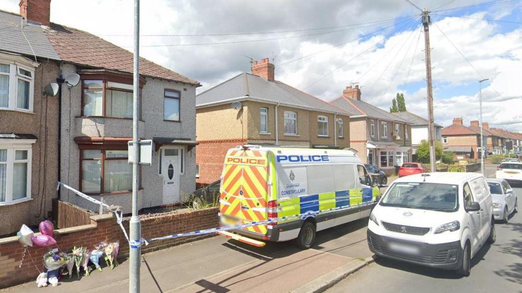 A police van parked outside a grey-rendered two-storey semidetached home on a street of houses. Blue and white police tape has been stretched around the house and about eight bouquets of flowers and some balloons have been left leaning against the small brick wall in front of the home.