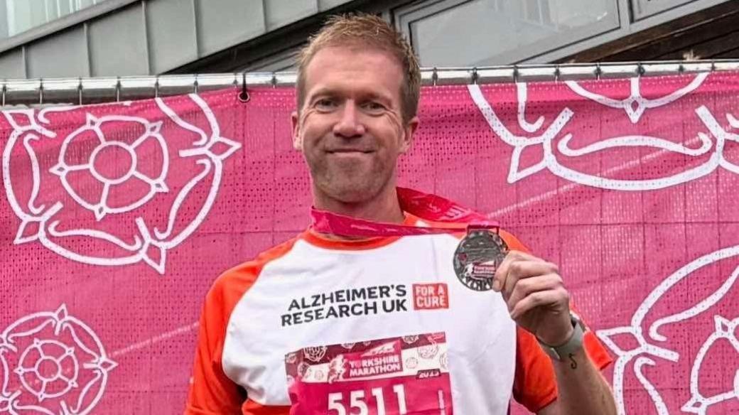 A man holds a silver model proudly, and smiles at the camera. He wears a white and orange t-shirt stating 'Alzheimer's Research UK', and has a pink bib for the Yorkshire Marathon with the number '5511' on. The man stands in front of a pink backdrop with white rose emblems on.