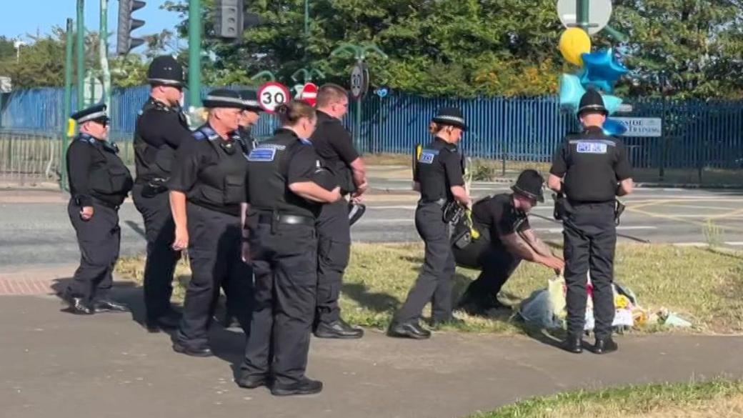 Eight police officers in uniform stand and watch as one of their colleagues puts down a floral tribute on an area of grass next to a road.