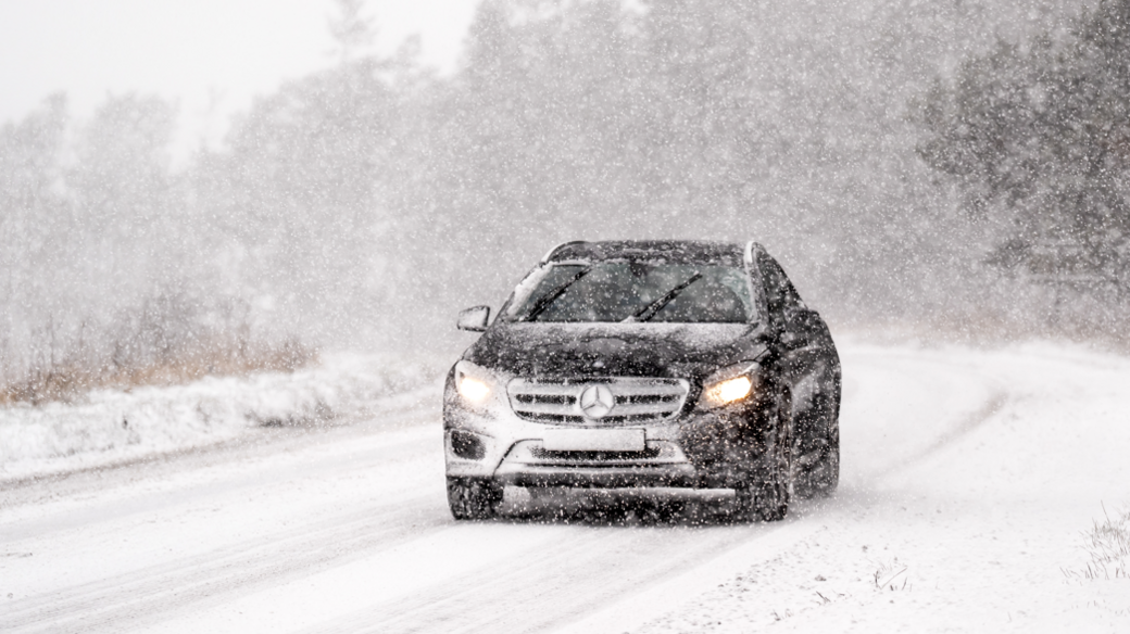 A black Mercedes Benz has its headlights and windscreen wipers on as it navigates through a snowstorm on a road which is already covered in snow in the North York Moors National Park