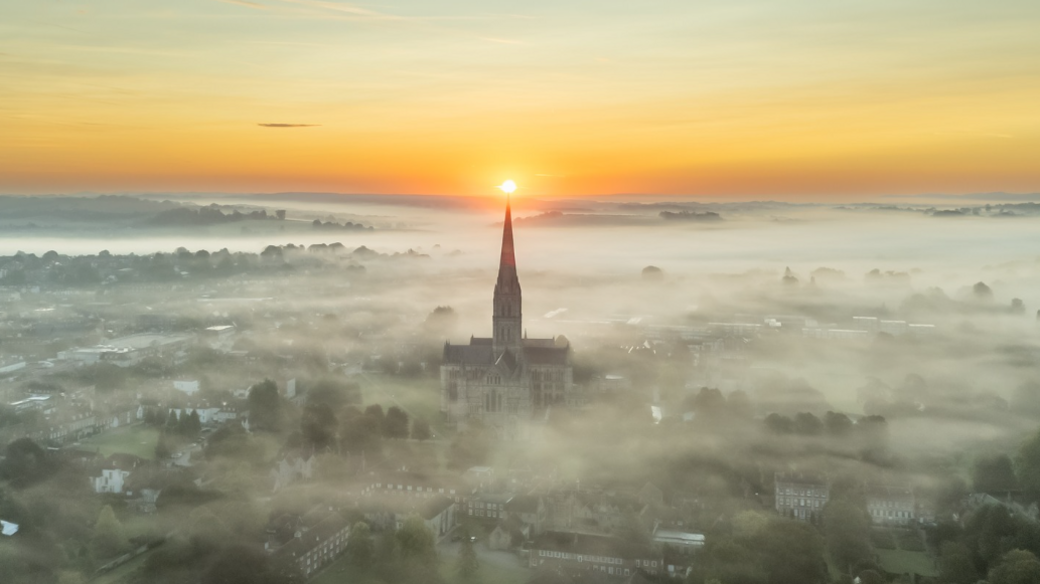 A drone shot of Salisbury Cathedral emerging from fog with the sun just rising on the horizon