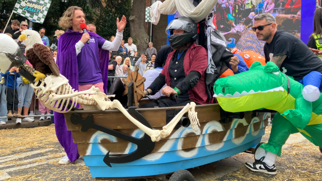 A boat shaped kart is on a road. Someone is sat in the kart which has blue and white waves painted on them. A person on the right is wearing an inflatable green dinosaur and pushing the kart.