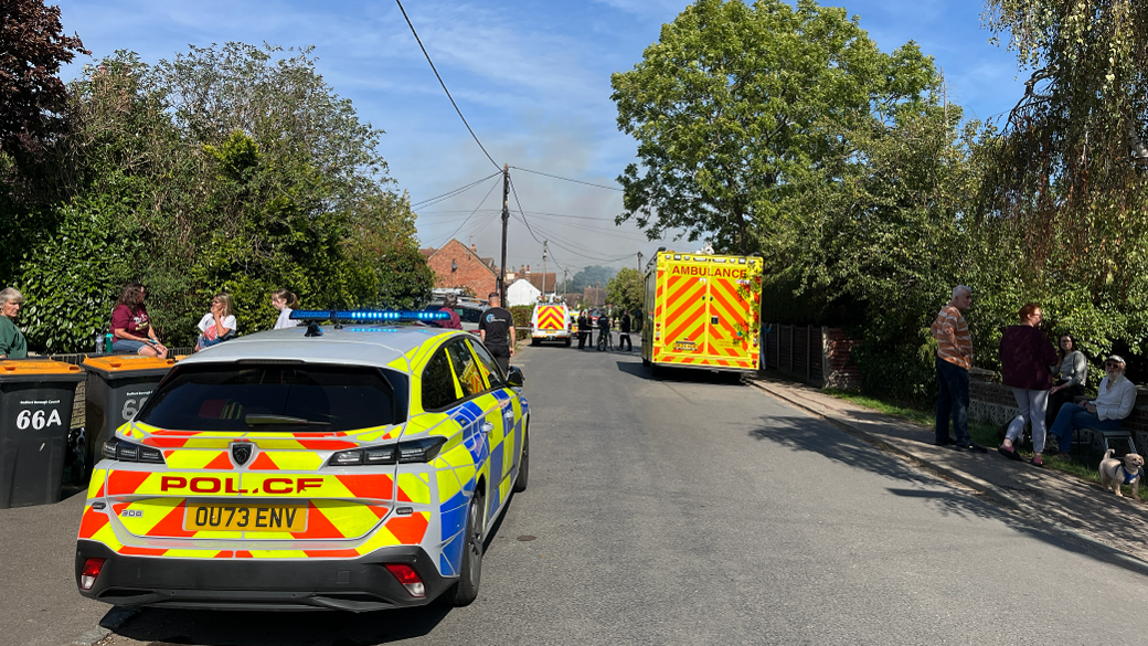Police emergency vehicle parked on a street with an ambulance parked further down the road. Residents are stood outside. There is some light smoke in the distance