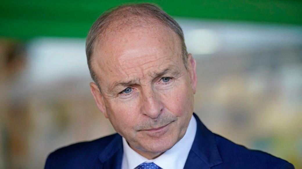 Micheál  Martin with balding light brown hair and blue eyes wearing a dark navy suit and looking towards someone speaking to him