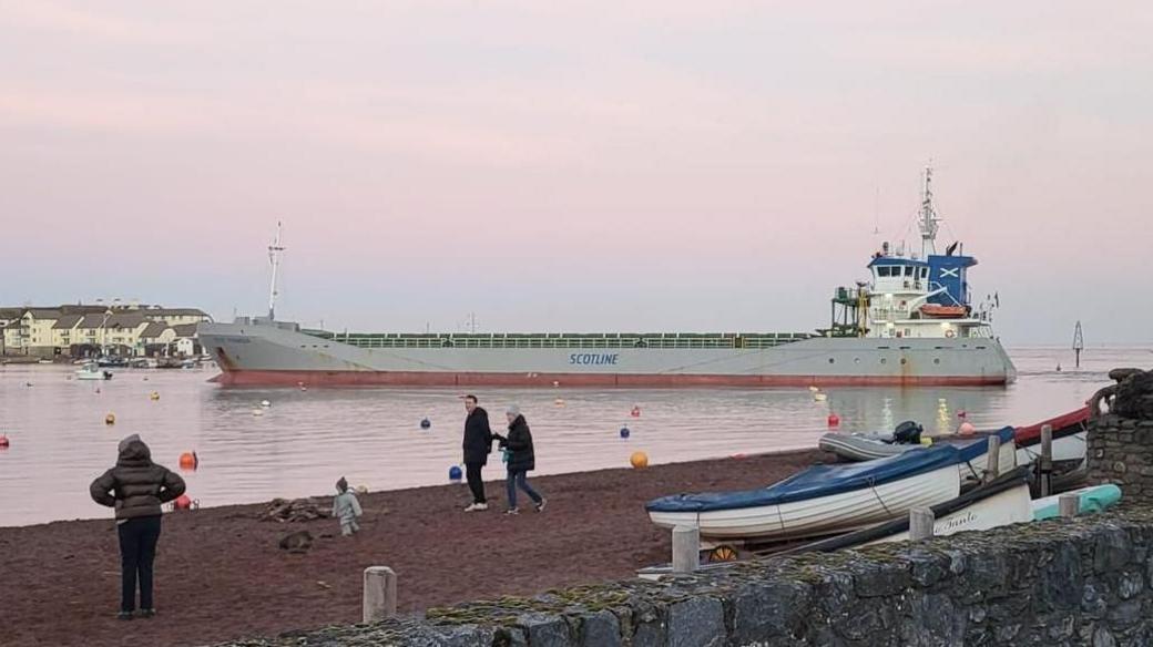 A large silver cargo ship called Scot Pioneer is seen in the water in Teignmouth Harbour. Three adults a small child are seen in the foreground on the beach at Shaldon.